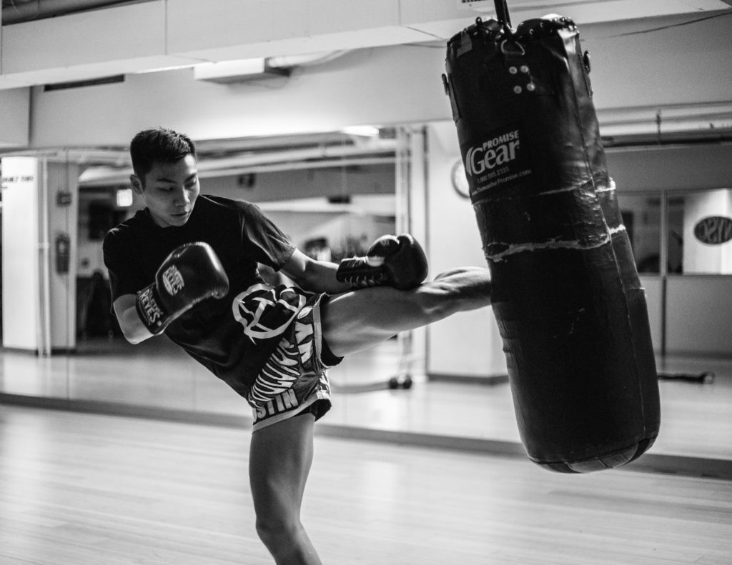 man wearing training gloves kicking heavy bag inside gym near mirror walls